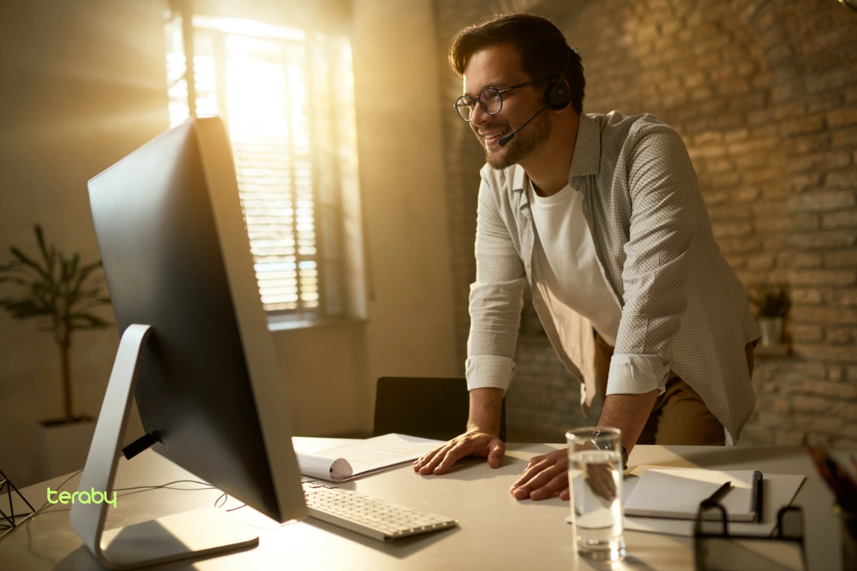 Hombre sonriente con audífonos y micrófono, apoyado en un escritorio frente a un monitor, en una oficina iluminada por el sol de la tarde